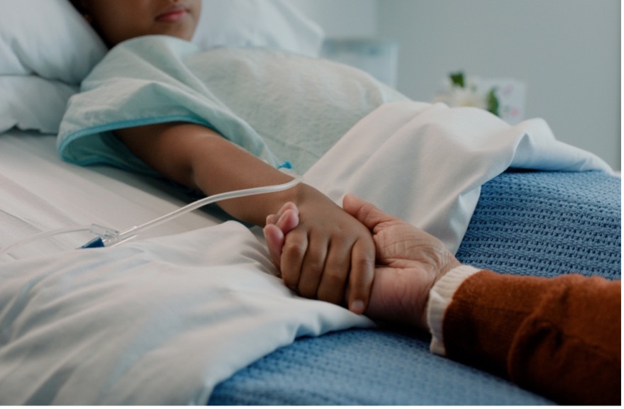 An adult holding the hand of a child resting in a hospital bed with an IV.