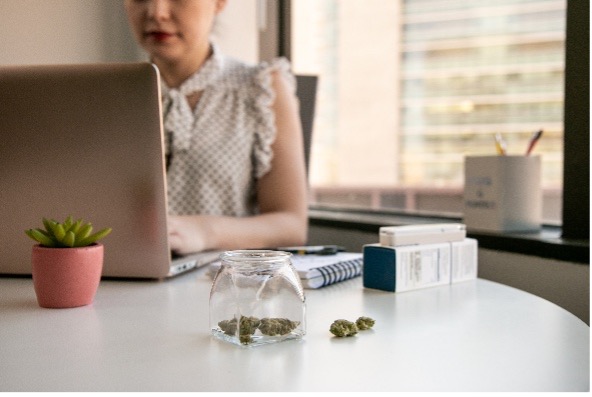 Cannabis buds in a glass jar on a white desk, with a woman working on a laptop in the background.